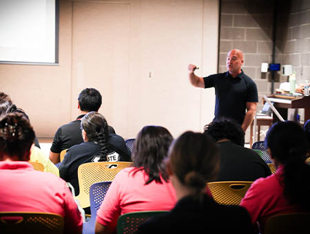 A presenter in a black shirt stands at the front of a room, gesturing with his hand while speaking to an audience seated in rows of colorful chairs. The view is from behind the audience, focusing on the speaker at the front.